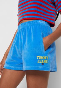 Modré sametové šortky s elastickým pasem. Mají boční kapsy a na levé noze je vyšívaný nápis "TOMMY JEANS" ve žluté barvě.