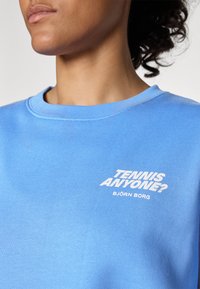 Sweatshirt azul claro feito de tecido macio, apresentando o texto branco "TENNIS ANYONE?" e "BJÖRN BORG" na parte da frente. Design clássico de gola redonda.