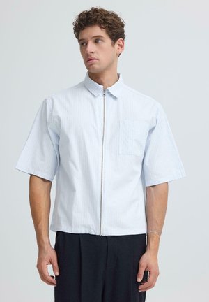 Ung mand med krøllet hår iført en lyseblå kortærmet skjorte med lynlås og mørke bukser, der kigger let til siden.