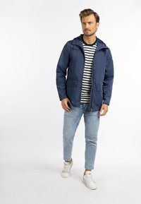 Marineblå udendørsjakke med hætte, lynlåslukning og to frontlommer, båret over en sort og hvid stribet skjorte og lyse denimjeans.