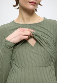 Žalia suknelė su briaunotu raštu, ilgomis rankovėmis, V formos iškirpte ir raukiniu krūtinės srityje. Minkšta, tekstūruota medžiaga su subtiliu blizgesiu.