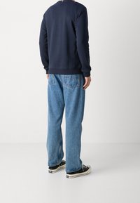 Felpa blu navy con polsini a costine, abbinata a jeans in denim azzurro a vestibilità comoda e sneaker nere. Design minimalista, senza motivi visibili.