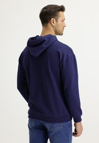 Felpa con cappuccio in cotone blu navy, maniche lunghe, polsini e orlo a coste, e vestibilità comoda. Dettagli di cucitura visibili alle cuciture delle spalle.