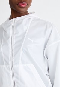 Biała lekka kurtka z kapturem, wyposażona w przedni zamek błyskawiczny i subtelne logo Puma. Gładka tekstura, minimalistyczny design o dopasowanym kroju.