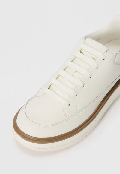 Minimalist kreemikatjaline spordijalats valgete paeltega ja pruuni äärega talla kohal, vastamisi lihtsa valge taustaga.