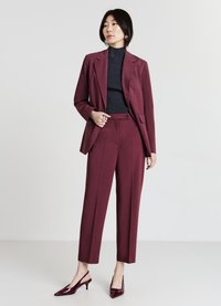 Burgunderfarbener maßgeschneiderter Anzug mit schmal geschnittenen Hosen und passendem Blazer, getragen über einem dunkelgrauen gerippten Rollkragenpullover; glänzende burgunderfarbene Absätze.