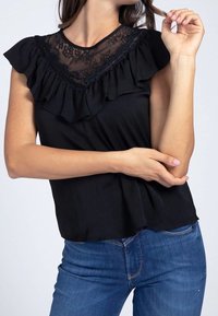 Top negro con escote de encaje, mangas rizadas, tela suave y corte relajado. Combinado con jeans azules, con un dobladillo curvo que se asoma.