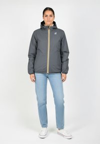 Grijze waterdichte jas met een gele rits, capuchon en elastische manchetten, gecombineerd met lichtblauwe jeans en witte sneakers. Eenvoudig ontwerp.