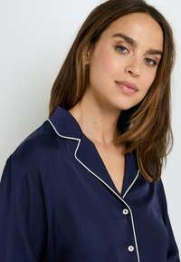 Haut de pyjama bleu marine en tissu doux avec passepoil blanc, présentant un col cranté, une fermeture à bouton et une coupe décontractée.