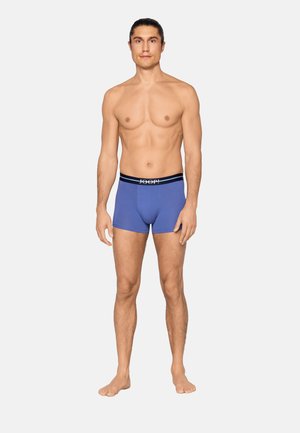 Młody mężczyzna stoi boso, zwrócony przodem, ubrany w niebieskie bokserki z czarnym paskiem w talii i białym logo na jednolitym tle.