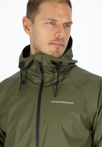 Grüne wasserdichte Jacke mit hohem Kragen, verstellbaren Kordelzügen und einem Frontreißverschluss. Verfügt über ein weißes Logo "SWEDEMOUNT" auf der Brust.