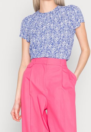 Femme portant un haut à manches courtes bleu et blanc à motif floral rentré dans un pantalon large taille haute rose vif, les mains dans les poches.