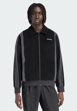 Črna kordjakna z sivim obrobjem, zadrgo in ovratnikom. Ima izšito modro Adidasovo logo na prsih; rebrasti manšeti.