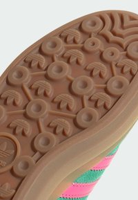Sneakersula med en texturerad gummiyta, som har cirkulära och bladliknande mönster i ljusbrunt, med ett överdel i grön och rosa tyg.