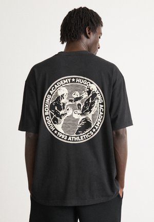 Vīrietis ģērbts melnā oversize T-kreklā ar apļa formas vintage boksa grafiku un tekstu "Hugo Boksa Akadēmija 1993 Atlētika" aizmugurē.