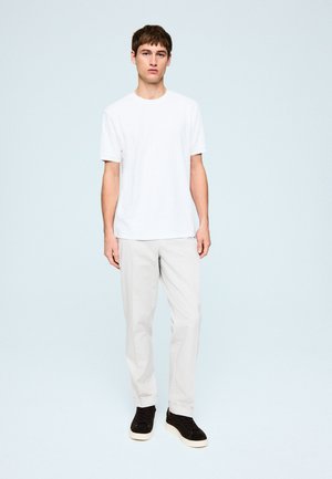 Junger Mann steht, trägt ein schlichtes weißes T-Shirt, hellgraue Hose und schwarze Sneakers mit weißen Sohlen.