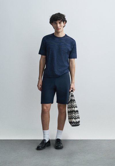 Μπλουζάκι με κοντά μανίκια και μπλε ναυτικό ριγέ σχέδιο, με στρογγυλή λαιμόκοψη, συνδυασμένο με σκούρα σορτς. Το μοντέλο κρατά μια τσάντα tote με ασπρόμαυρο σχέδιο Chevron.