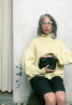 Gele oversized coltrui, zwarte geweven handtas, zittend op een gestructureerd oppervlak, neutrale achtergrond met gedrapeerde stof.