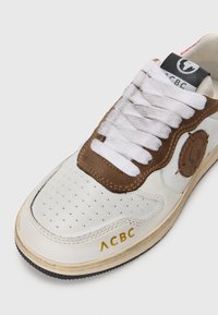 Valged spordijalatsid perforeeritud disainiga, pruun nahkdetailide ja valgete nööpidega. Külgedel on logo silt ja kuldne "ACBC" kiri.