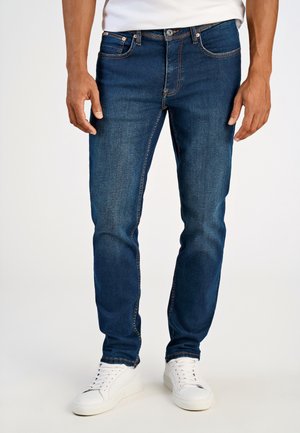Mann trägt dunkelblaue Slim-Fit-Jeans und weiße Sneakers, steht vor einem schlichten hellen Hintergrund.
