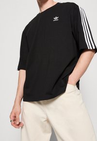 Svart oversized t-shirt med rund halsringning, tre vita ränder på ärmarna och en liten vit logotyp på bröstet.