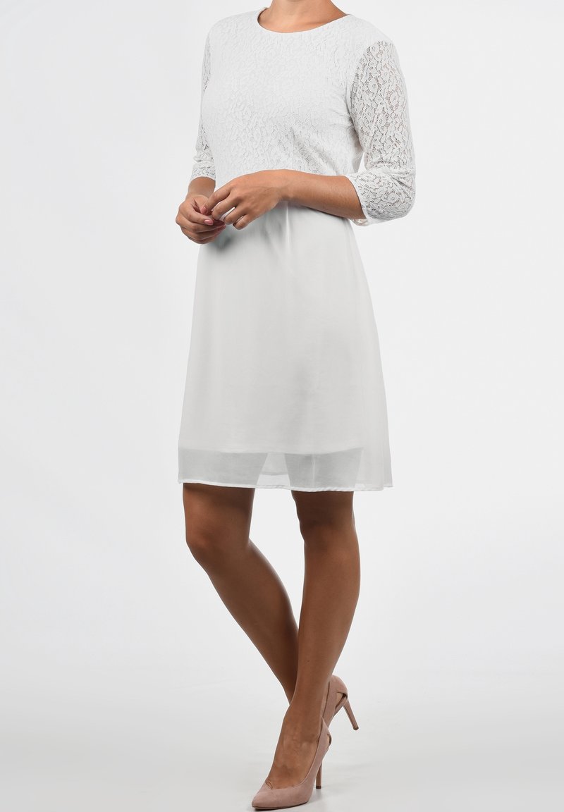 Biała sukienka z koronkowymi rękawami do połowy ramienia i dopasowanym gorsetem. Zwiewna spódnica z szyfonu o długości do kolan, w połączeniu z jasnoróżowymi szpilkami.