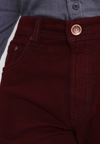 Pantaloni in denim bordeaux con un bottone in rame e cuciture scure; un classico modello a cinque tasche con un tessuto leggermente strutturato.