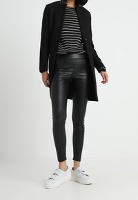 Svart lång jacka över en svartvit randig topp, kombinerad med glänsande svarta leggings och vita sneakers. Enkel design, modern passform.