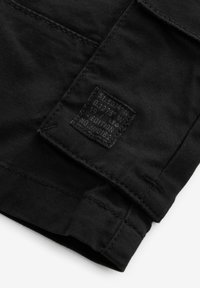 Tissu noir avec une étiquette texturée comportant du texte blanc. L'étiquette contient des coordonnées, des détails d'édition et un design en relief.