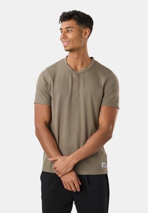 Giovane uomo con capelli corti e ricci che indossa una maglietta henley oliva a maniche corte e pantaloni neri, che guarda di lato con le mani intrecciate.