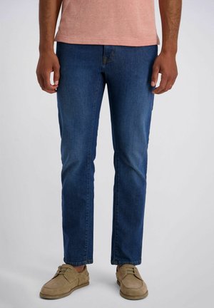 Mann trägt enge blaue Jeans, ein hellrosa Hemd und beige Schnürschuhe, steht vor einem schlichten weißen Hintergrund.