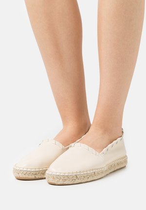 Pieds portant des espadrilles en cuir de couleur crème avec des semelles en jute tressé et des détails en surjet autour des bords sur un fond blanc.
