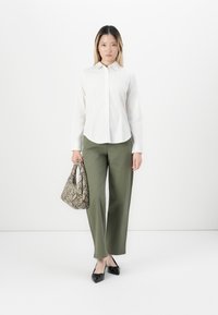 Woman standing against a white background wearing a white button-up shirt, olive green pants, black shoes, and holding a snakeskin-patterned handbag.