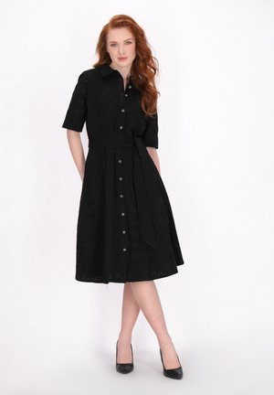 Schwarzes Knopfkleid mit kurzen Ärmeln, versehen mit Lochstickerei, tailliertem Schnitt und einem passenden Gürtel. Kombiniert mit schwarzen Absatzschuhen.