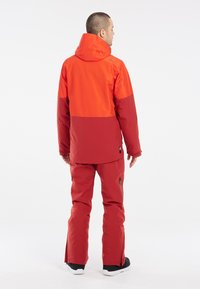 Rote und orange Skijacke mit Kapuze und kontrastierenden Farbblöcken, kombiniert mit passenden roten Hosen mit Reißverschlusstaschen und schwarzen Akzenten.