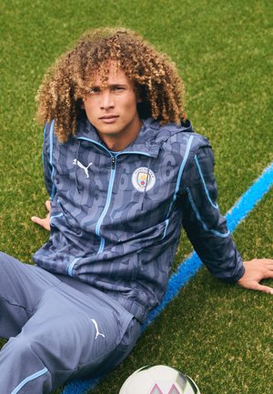 Sudadera Puma en gris oscuro con detalles en azul, patrones geométricos y el logo del Manchester City; combinada con pantalones gris claro y un balón de fútbol.