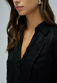 Czarna bluzka z kwiatowym wzorem, teksturowany materiał i wyraźnym kołnierzykiem. Projekt zawiera subtelne pionowe paski oraz głęboki dekolt.