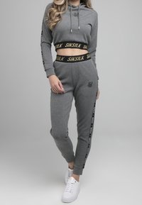 Grijze cropped hoodie met trekkoorden en logo, gecombineerd met bijpassende joggers met zijkstrepen en een zwarte tailleband met tekst. Witte sneakers.