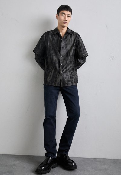Camisa negra de manga corta con un acabado texturizado y sutiles rayas, combinada con jeans ajustados de color azul oscuro y zapatos negros con cordones.