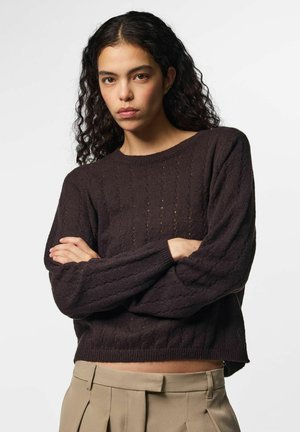 Jeune femme aux cheveux bouclés foncés portant un pull en maille marron foncé et un pantalon beige taille haute, les bras croisés, expression neutre.