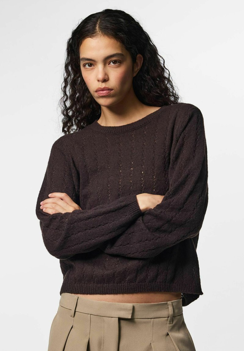 Jeune femme aux cheveux bouclés foncés portant un pull en maille marron foncé et un pantalon beige taille haute, les bras croisés, expression neutre.