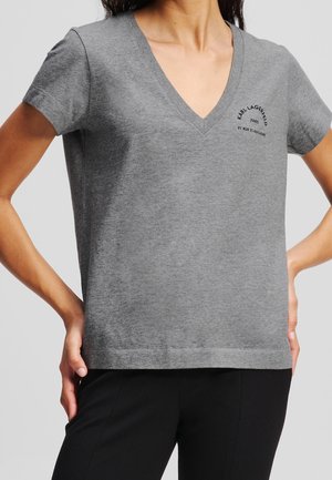 Kvinde iført en grå V-hals T-shirt med "Karl Lagerfeld Paris 21 Rue St-Guillaume" logo på venstre bryst, kombineret med sorte bukser.