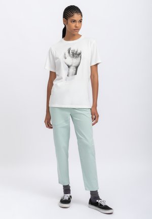 T-shirt bianca in cotone con una grafica nera di una mano alzata; abbinata a pantaloni verde menta e sneakers nere con suole bianche.