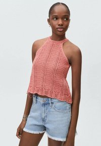 Häkelad halterneck-top i mjuk korall, med en texturerad mönster och vågig nederkant. Kombineras med ljusblå denimshorts med fransade kanter.