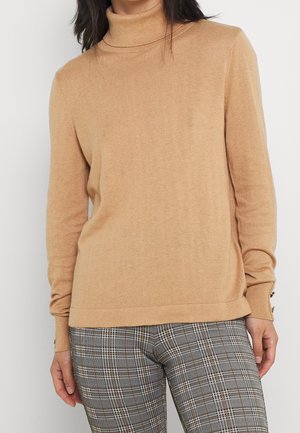 Pull à col roulé beige clair en tricot doux, avec des manches longues et des accents de boutons subtils sur les poignets. Associé à un pantalon à carreaux.