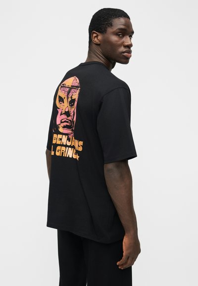 Mężczyzna noszący czarny, oversize'owy t-shirt z różowo-pomarańczową grafiką maski lucha libre i napisem "Benjamins El Gringo" na plecach.