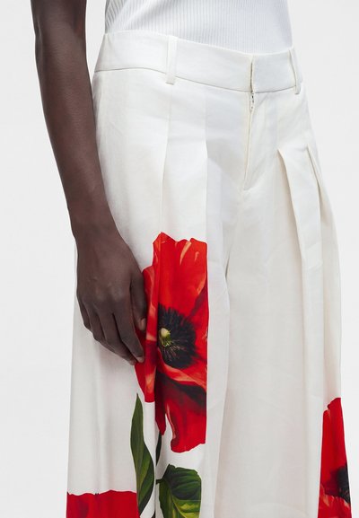 Gros plan d'une personne portant un pantalon blanc avec de grandes fleurs de coquelicot rouges imprimées et un haut blanc côtelé, montrant une partie de son bras à la peau foncée.