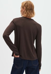 Person mit kurzen blonden Haaren, die ein dunkelbraunes Langarmshirt und dunkelblaue Jeans trägt, von hinten vor einem einfarbigen Hintergrund gesehen.