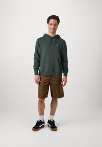 Green hooded sweatshirt with front pouch pocket, paired with brown shorts and black sneakers, featuring white accents and a textured fabric.