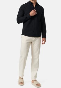 Czarna koszula z długim rękawem i kołnierzykiem na guziki, zestawiona z beżowymi spodniami o fakturze i jasnobeżowymi klapkami na stopę.